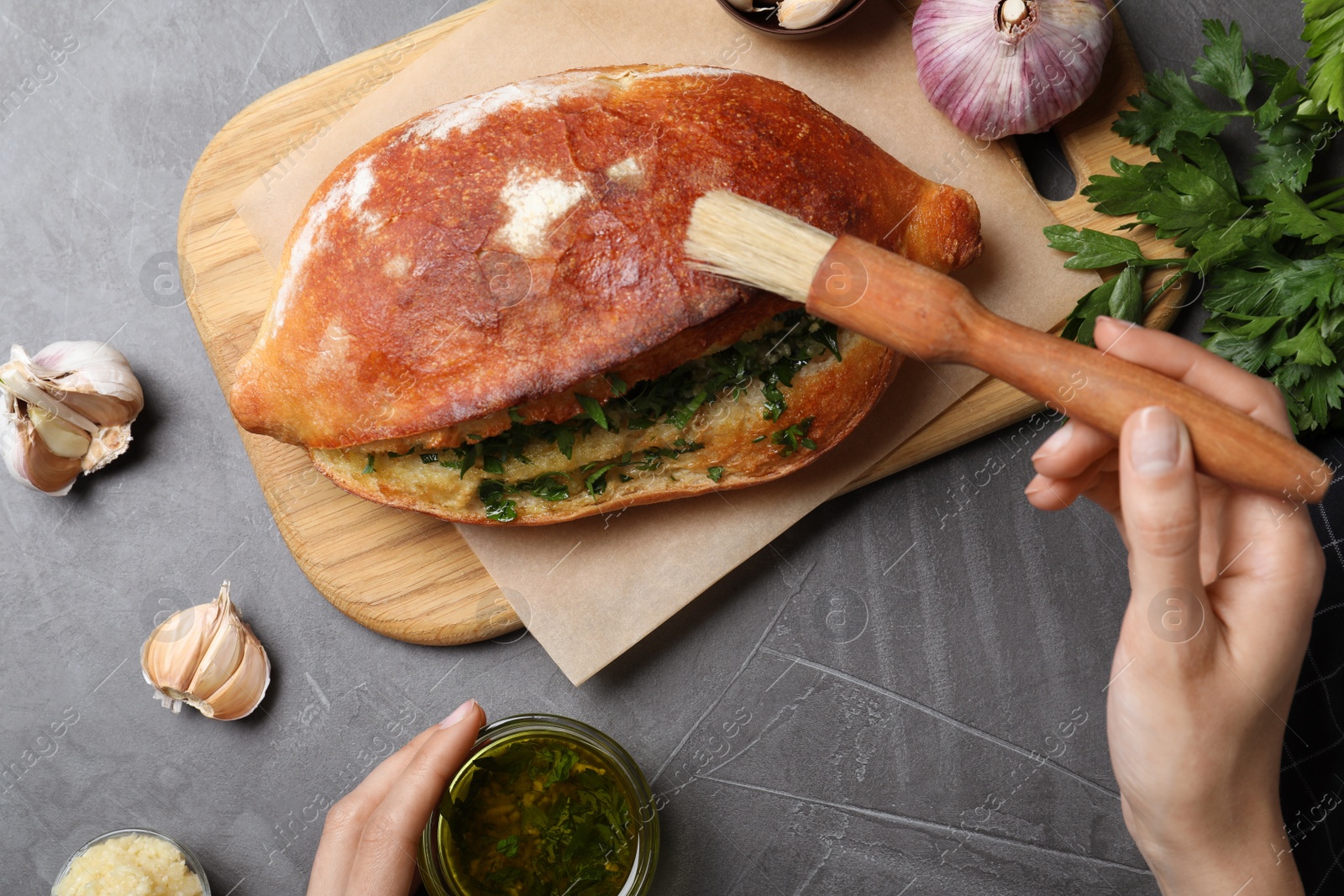 Photo of Woman brushing delicious homemade bread with herbs and garlic at grey table, top view