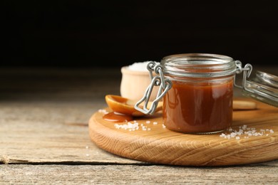 Tasty salted caramel in jar and salt on wooden table, space for text