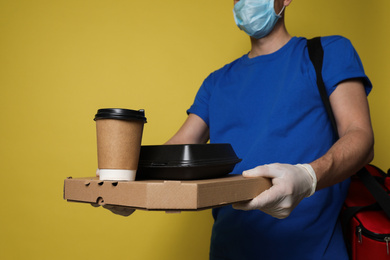 Courier in protective mask and gloves holding order on yellow background, closeup. Food delivery service during coronavirus quarantine