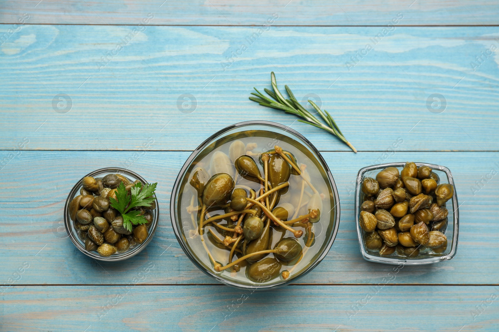 Photo of Tasty capers, rosemary and parsley on light blue wooden table, flat lay