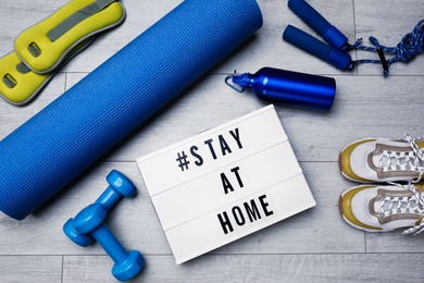 Photo of Sport equipment and lightbox with hashtag STAY AT HOME on floor, flat lay. Message to promote self-isolation during COVID‑19 pandemic