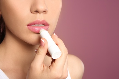 Young woman applying balm on her lips against color background, closeup