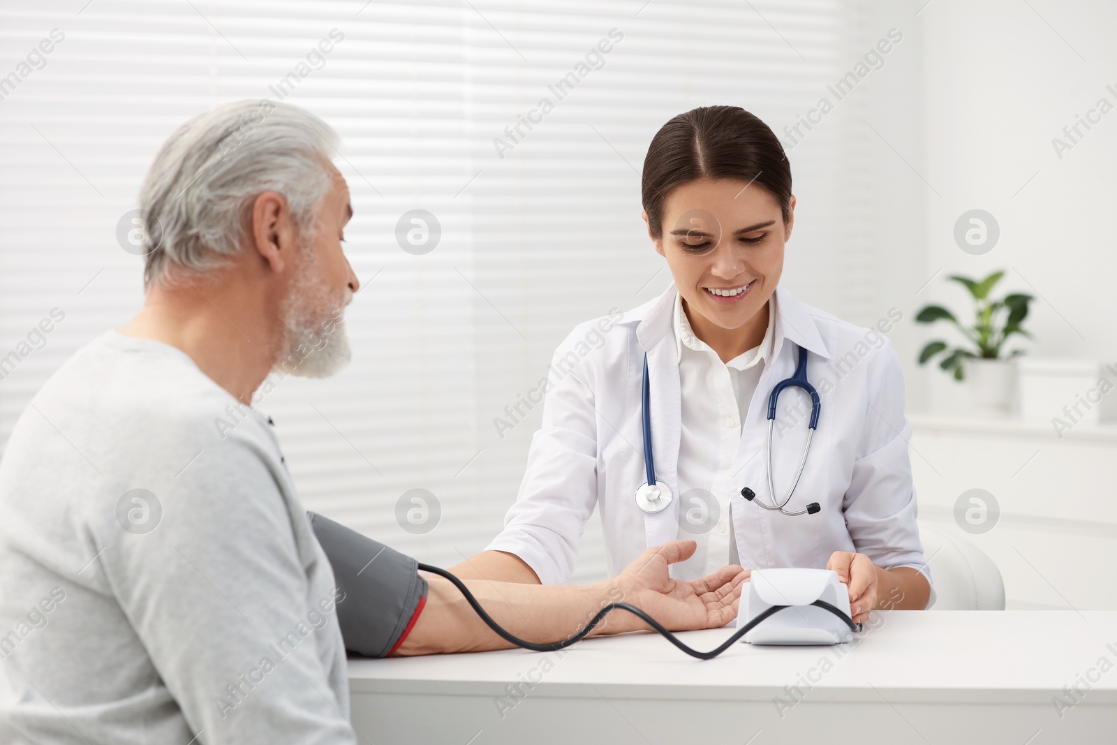 Photo of Smiling nurse measuring elderly patient's blood pressure at white table in hospital