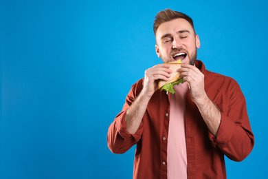 Young man eating tasty sandwich on light blue background. Space for text