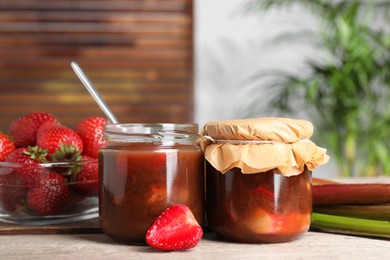 Jars of tasty rhubarb jam, fresh stems and strawberries on wooden table. Space for text