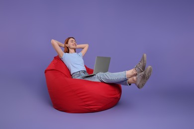 Happy young woman with laptop sitting on beanbag chair against lilac background