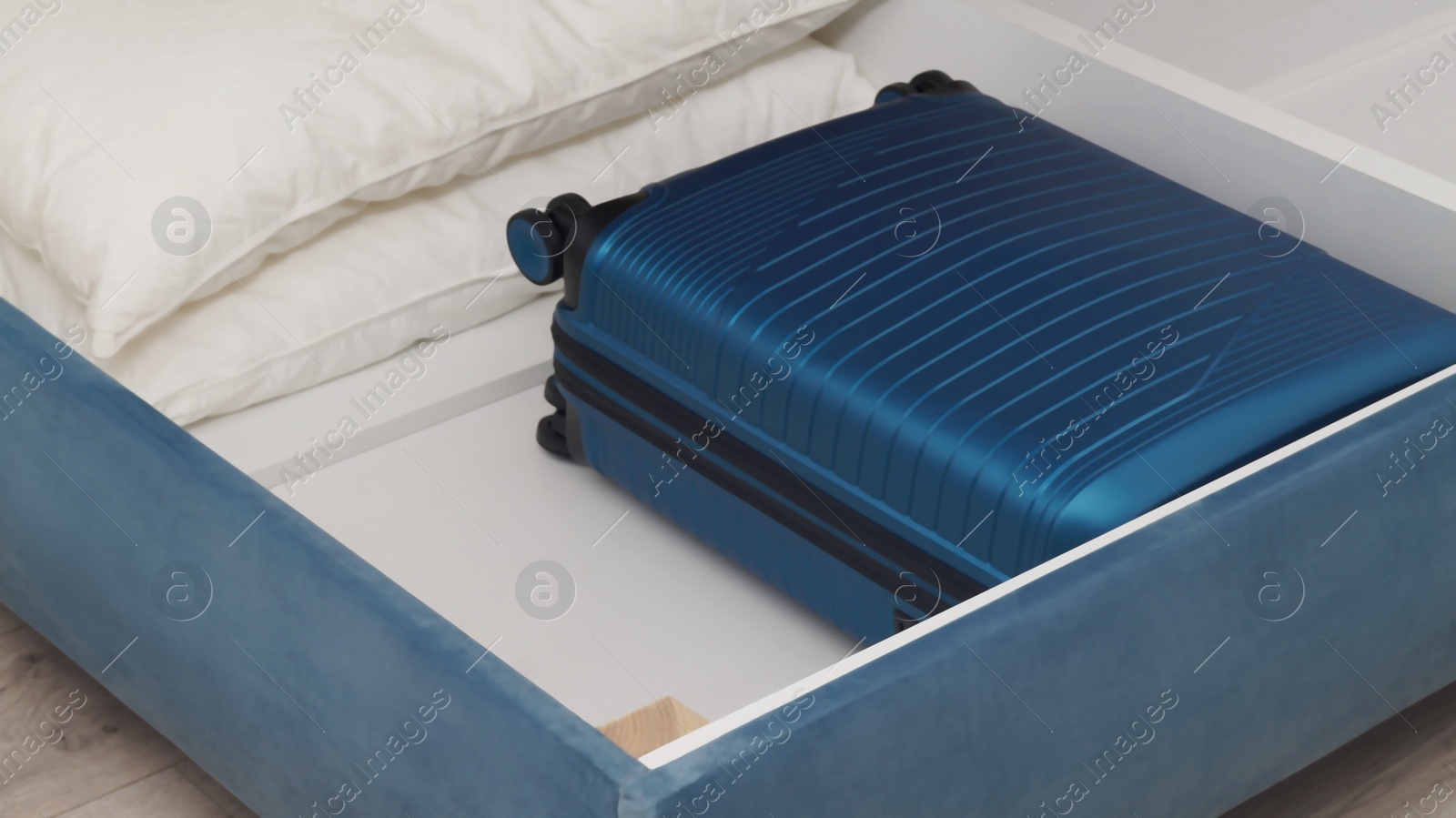 Photo of Storage drawer under bed with blue suitcase and white pillows indoors, closeup
