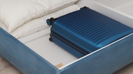 Storage drawer under bed with blue suitcase and white pillows indoors, closeup