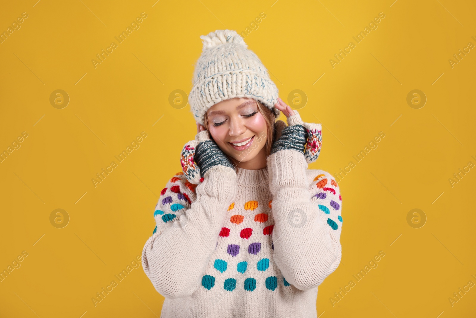 Photo of Young woman in warm sweater, mittens and hat on yellow background. Winter season