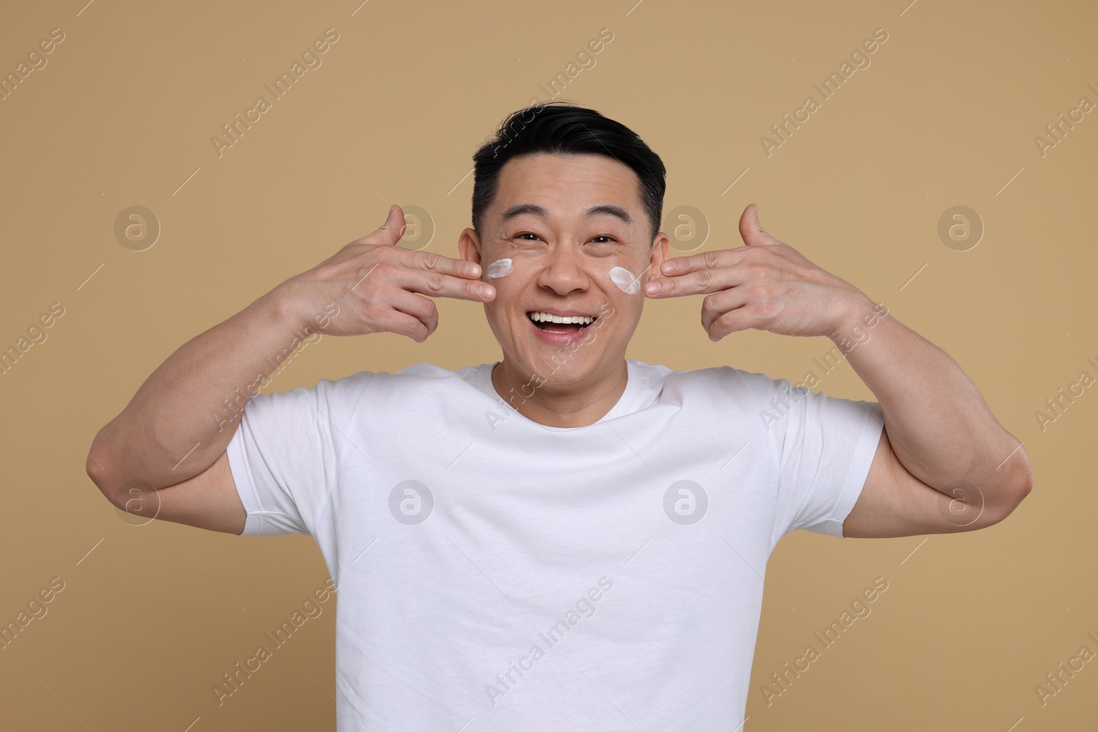 Photo of Emotional man applying cream onto his face on light brown background