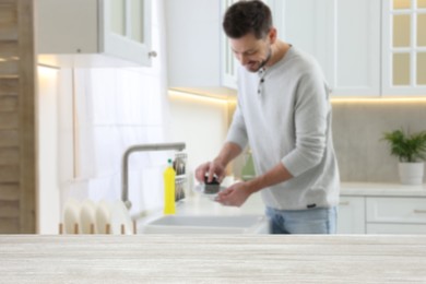 Image of Man doing washing up in kitchen, focus on empty wooden table