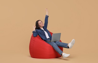 Photo of Happy young woman with laptop sitting on beanbag chair against beige background