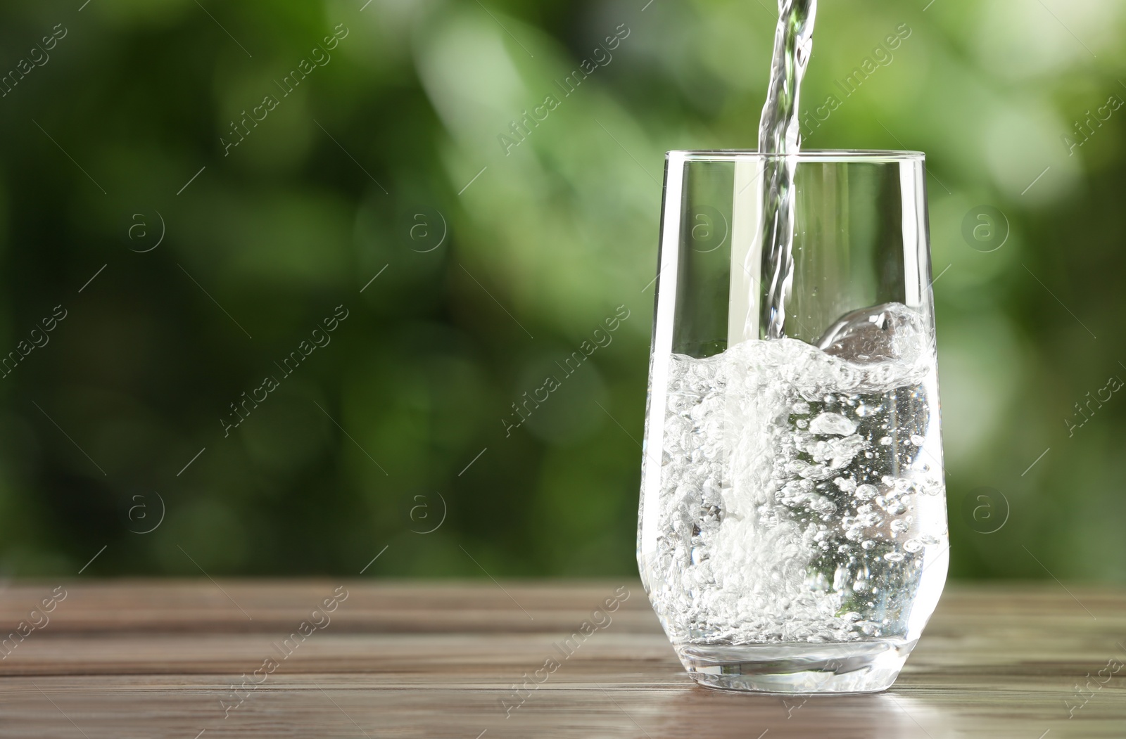 Photo of Pouring water into glass on wooden table outdoors, space for text