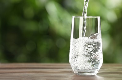 Photo of Pouring water into glass on wooden table outdoors, space for text