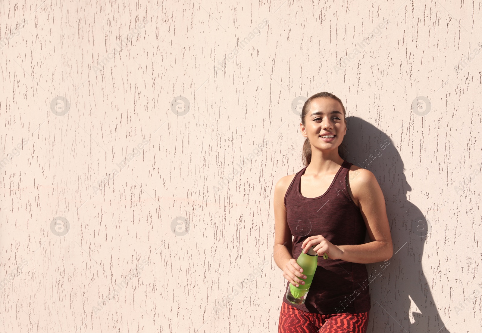 Photo of Young sporty woman holding bottle of water near wall outdoors on sunny day. Space for text