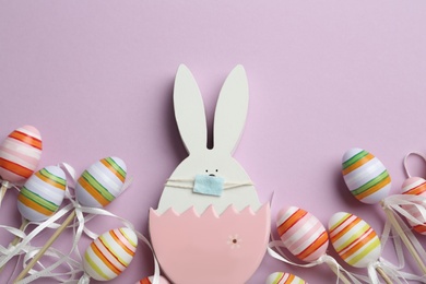 Photo of Wooden bunny with protective mask and eggs on lilac background, flat lay. Easter holiday during COVID-19 quarantine