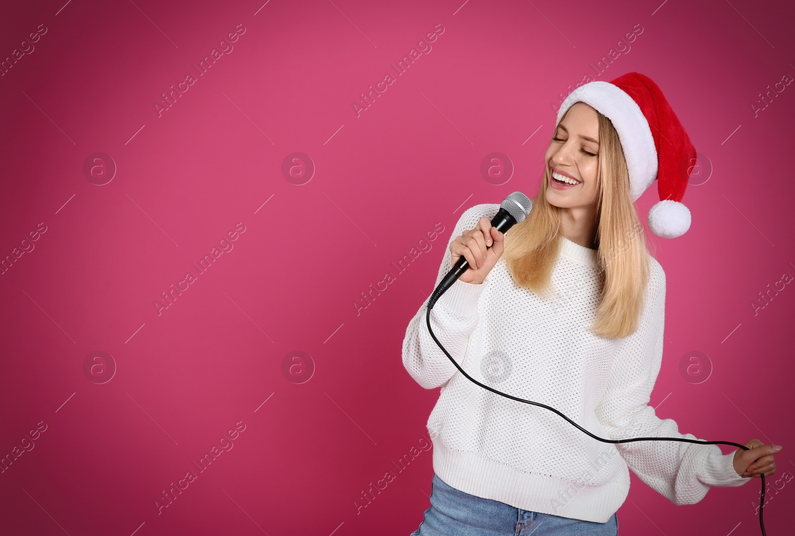 Photo of Happy woman in Santa Claus hat singing with microphone on pink background, space for text. Christmas music