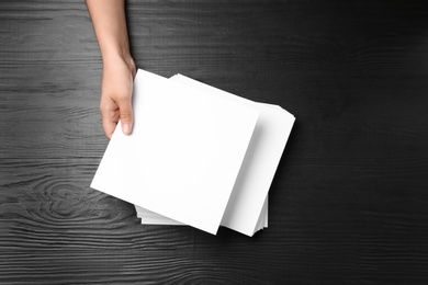 Woman holding blank paper sheets for brochure at black wooden table, top view. Mock up