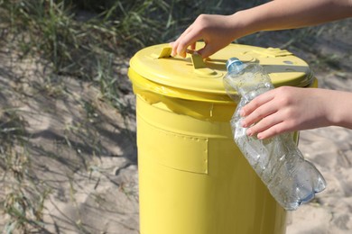 Photo of Woman throwing plastic bottle in yellow bin on beach, closeup. Recycling concept