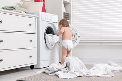 Photo of Little girl pulling baby clothes out of washing machine indoors