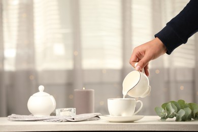 Woman pouring milk into cup at table indoors, closeup