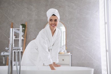 Photo of Beautiful young woman in soft white robe near bathtub at home