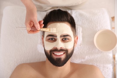 Photo of Cosmetologist applying mask on man's face in spa salon, top view