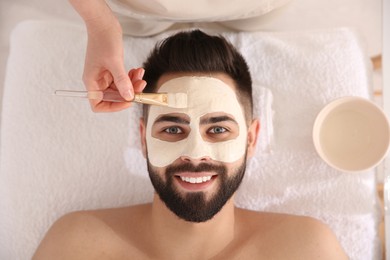 Cosmetologist applying mask on man's face in spa salon, top view