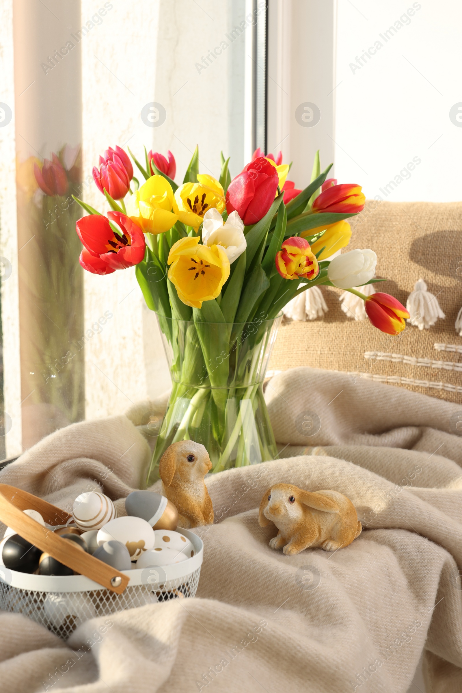 Photo of Easter decorations. Beautiful tulips, basket of painted eggs, bunny figures, pillow and plaid on window sill