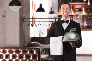 Photo of Waiter in elegant uniform holding metal tray and cloche at workplace