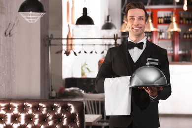 Waiter in elegant uniform holding metal tray and cloche at workplace