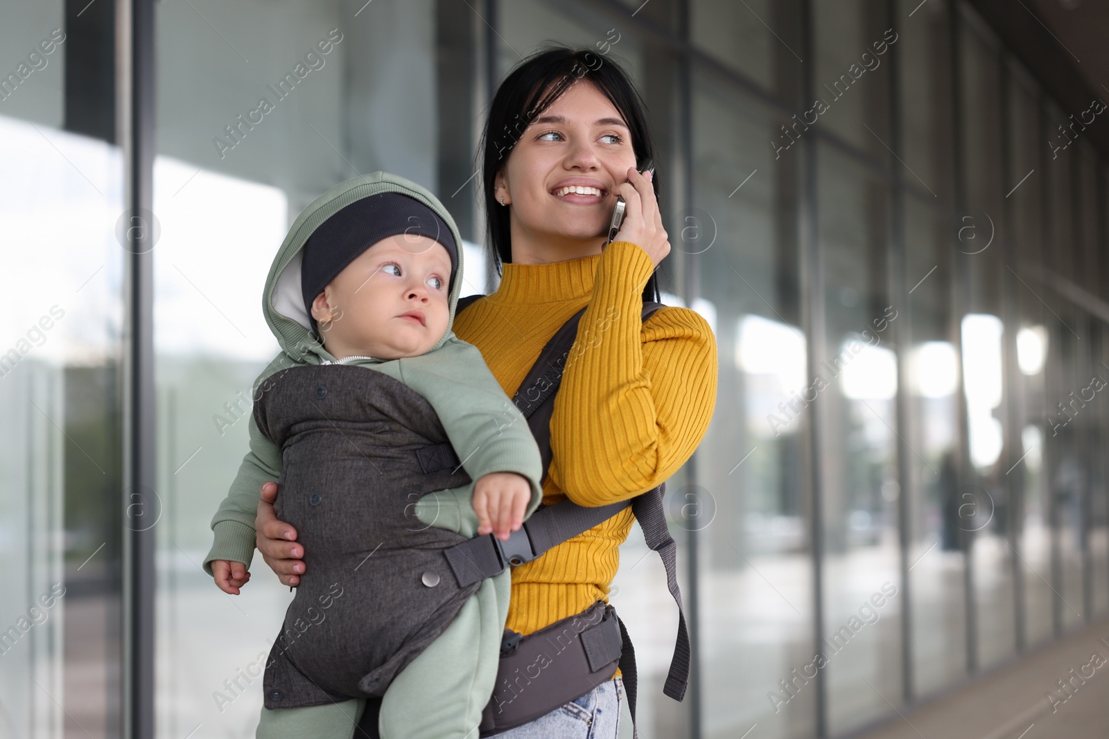 Photo of Mother holding her child in sling (baby carrier) while talking on smartphone near building outdoors