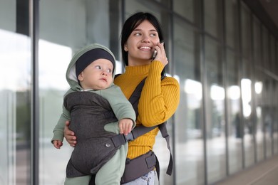 Mother holding her child in sling (baby carrier) while talking on smartphone near building outdoors