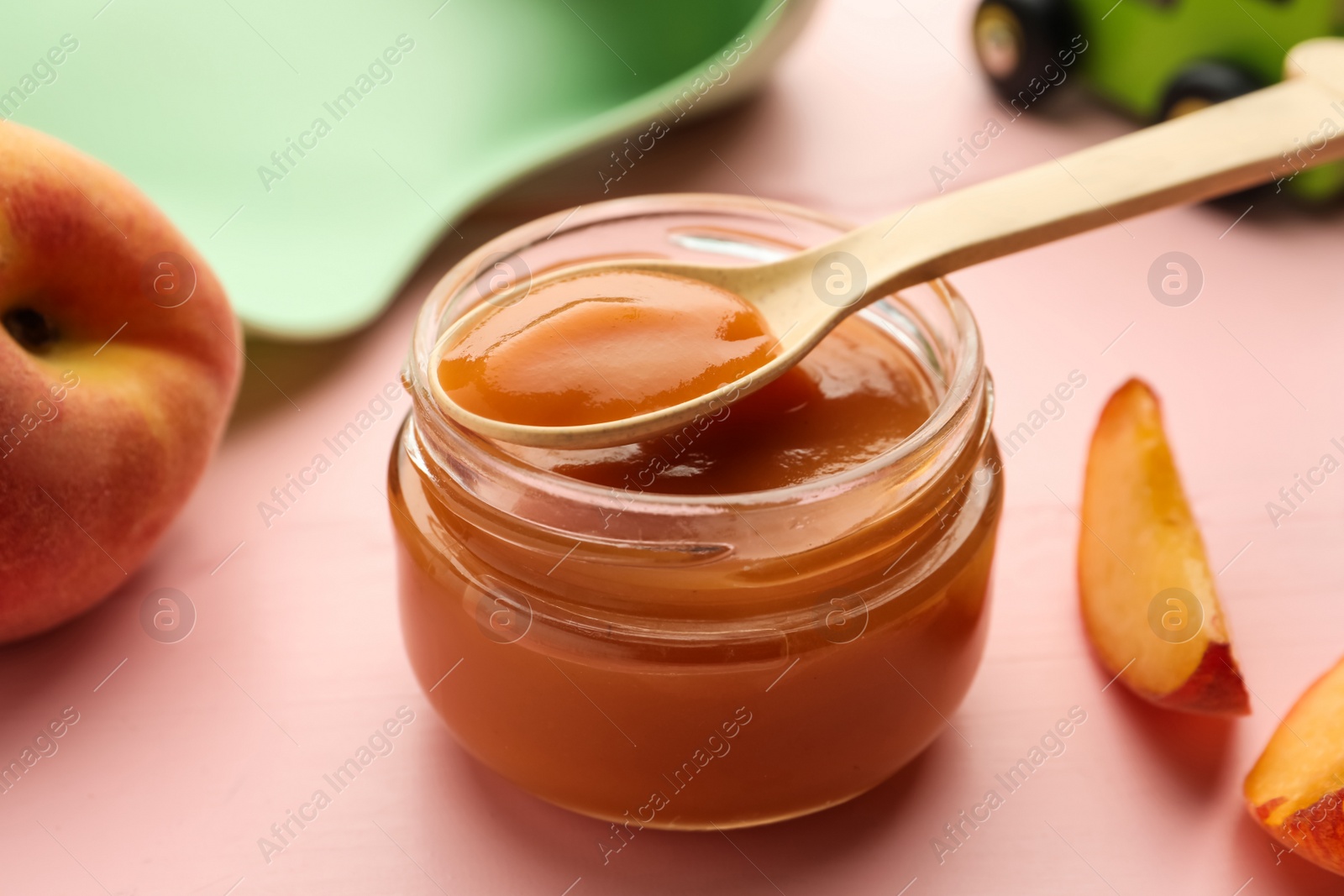 Photo of Spoon with healthy baby food over glass jar on pink wooden table, closeup