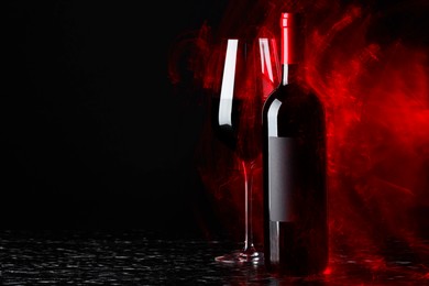 Tasty wine in glass and bottle in red lights on textured table against black background, space for text