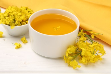 Photo of Rapeseed oil in bowl and beautiful yellow flowers on white wooden table