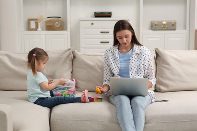 Little daughter playing while her mother working remotely on sofa at home