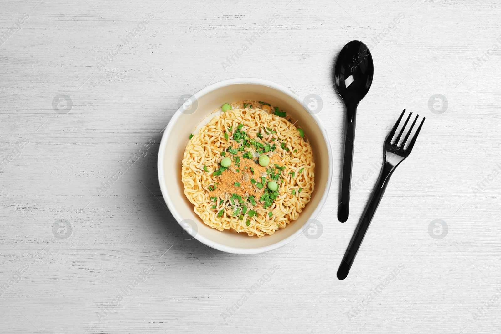 Photo of Cup of instant noodles with plastic cutlery on white wooden background, flat lay