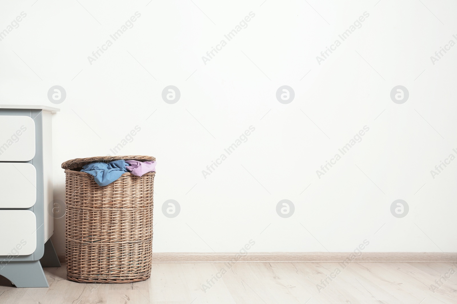 Photo of Wicker laundry basket with dirty clothes on floor near wall. Space for text