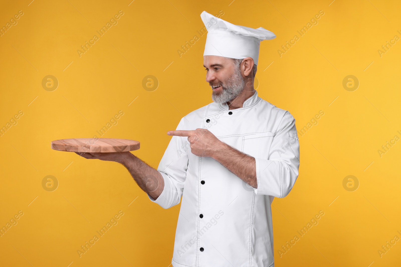 Photo of Happy chef in uniform pointing at wooden board on orange background