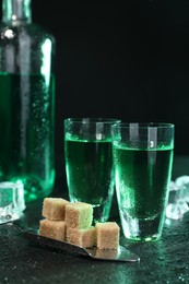 Photo of Absinthe in shot glasses, brown sugar cubes and spoon on gray table against dark background. Alcoholic drink