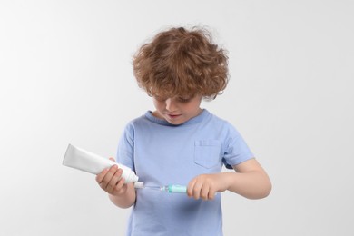 Cute little boy squeezing toothpaste from tube onto electric toothbrush on white background