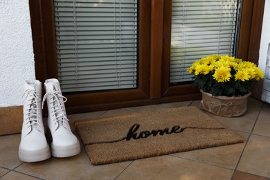 Doormat with word Home, flowers and stylish boots near entrance outdoors