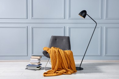 Comfortable armchair with blanket, books and lamp near grey wall indoors