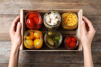 Photo of Woman holding crate with jars of pickled vegetables on wooden table, top view