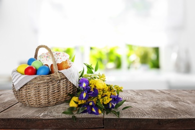 Basket with traditional Easter cake, dyed eggs and flowers on wooden table indoors. Space for text