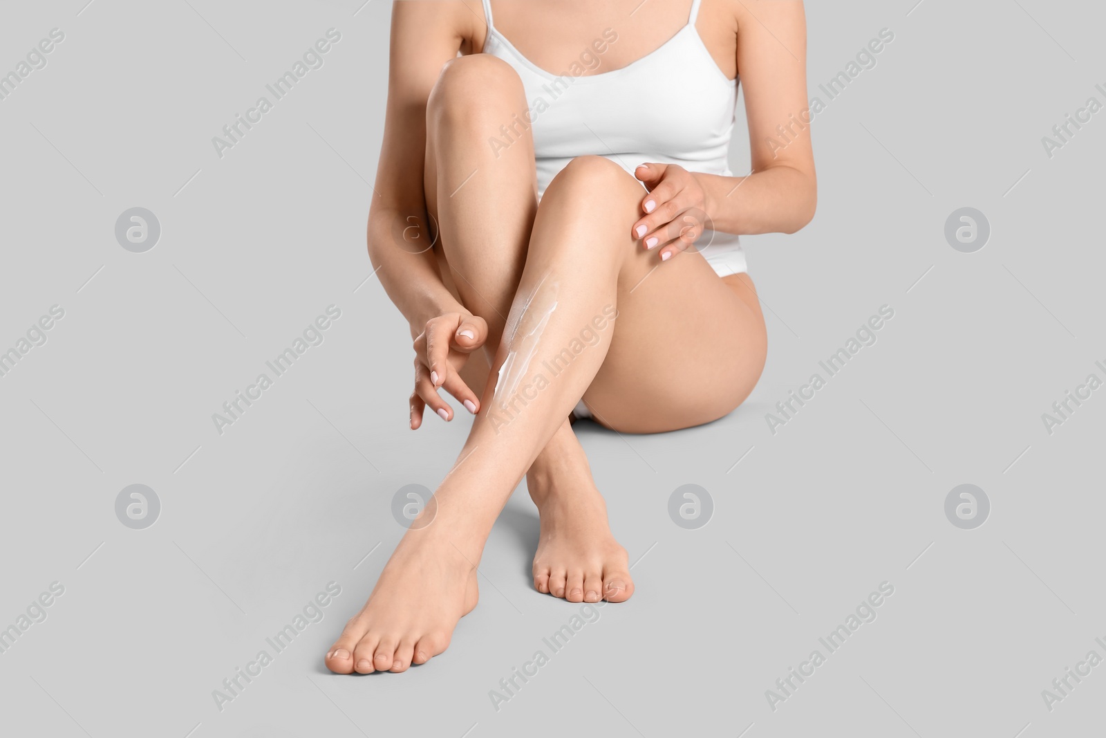 Photo of Woman applying body cream onto her smooth legs on light grey background, closeup