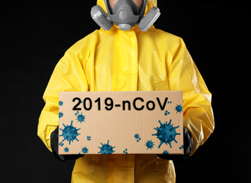 Man wearing chemical protective suit with cardboard box on black background, closeup. Coronavirus outbreak