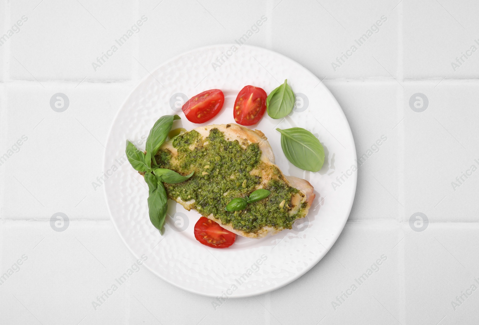 Photo of Delicious chicken breast with pesto sauce, tomatoes and basil on white tiled table, top view