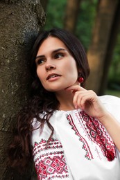 Photo of Beautiful woman in embroidered shirt near tree outdoors. Ukrainian national clothes